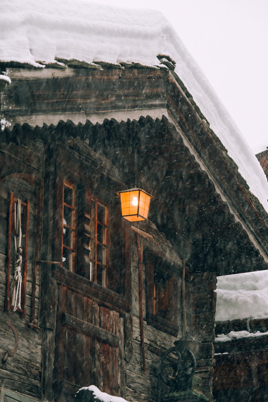 wooden cottage with burning lantern during snowfall